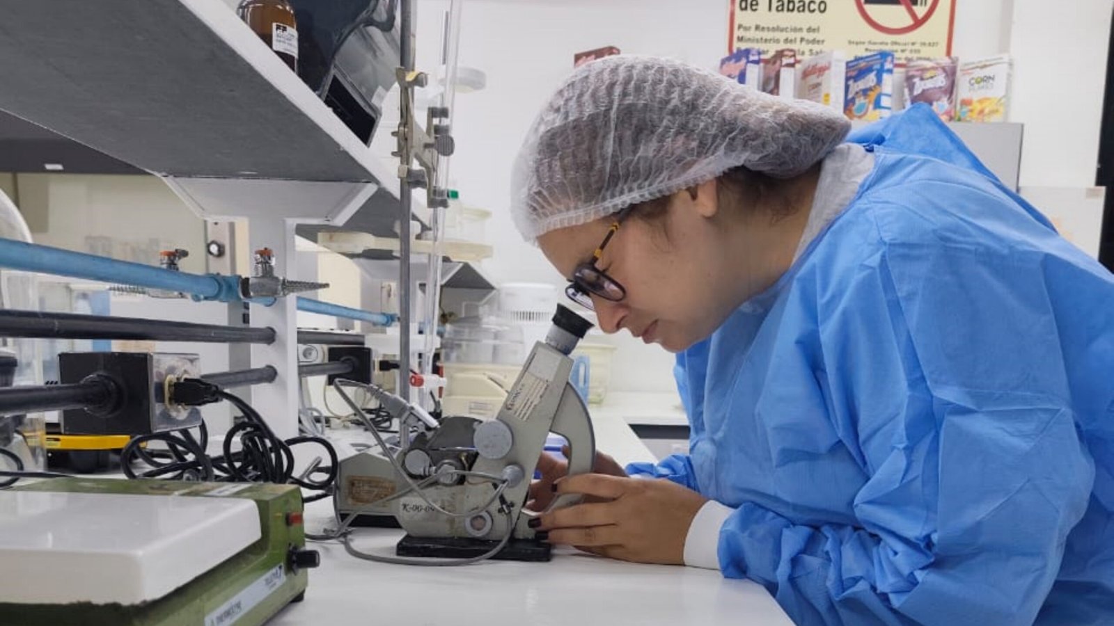 Norka Rojas con cofia y bata de laboratorio, trabajando en un microscopio