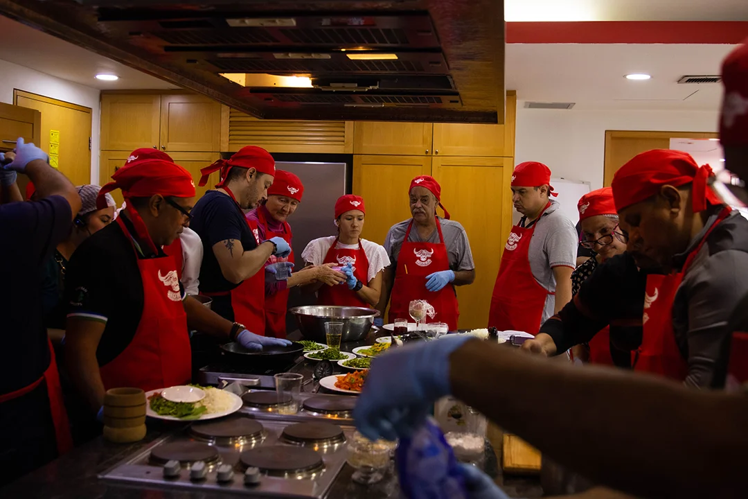 Grupo en una clase práctica de charcutería, con gorros y delantales rojos, alrededor de una mesa