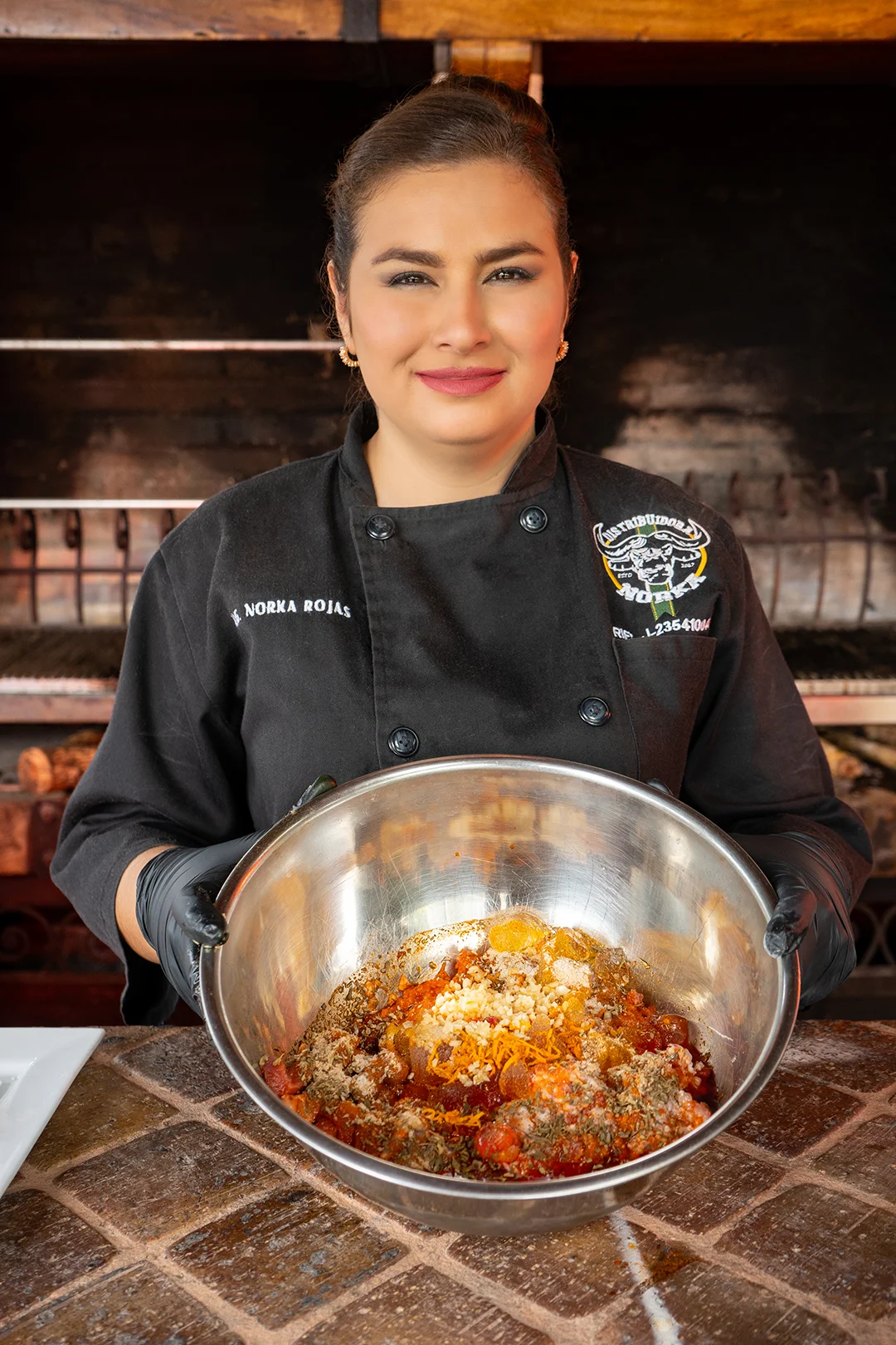 Norka Rojas en uniforme de chef sosteniendo un tazón metálico con mezcla de embutido, en la cocina